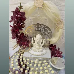 A wedding hamper designed to invoke blessings and joy. It features a serene Ganesha idol as the centerpiece, surrounded by a platter of sweets and adorned with rich, deep red orchids.