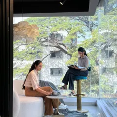 The front display area of the showroom, viewed from inside. The floor-to-ceiling glass wall floods the space with natural light and connects the luxurious interior with the trees outside, creating a serene and upscale atmosphere.