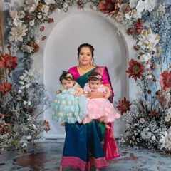 A proud mother holds her twin daughters in this beautiful portrait. The intricate floral backdrop complements her traditional saree, making for a truly artistic and heartfelt image.