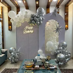 A modern and chic birthday setup in a living room. This design features elegant arched backdrops, a neon sign, and balloon clusters in a sophisticated silver and white color scheme.