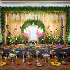 A clear, full view of a traditional stage decoration. The design is centered around a white Maharaja chair against a lotus-themed backdrop, framed by greenery and floral arrangements, with all the ceremonial trays neatly presented.