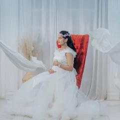 An elegant indoor studio setup with a red throne chair and flowing white fabrics. This creates a high-fashion, angelic look for a solo maternity portrait.