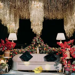 A breathtaking seating area for the happy couple, set under a cascading ceiling of gold tassels and crystal chandeliers. The combination of lush red florals and soft lamp lighting creates an atmosphere of pure romance and opulence.