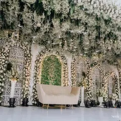 A closer view of our intricate green and white stage decor. The setup features beautiful white latticework panels, arches filled with greenery, and a ceiling overflowing with baby's breath and hanging glass birds.