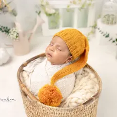 Wrapped snugly in a white swaddle and wearing a cute pom-pom hat, this baby is sleeping in a simple wicker basket. The natural textures create a warm and cozy feel.