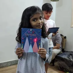 A young girl proudly shows off the light-up Christmas card she made. This project combines crafting with simple electronics, teaching kids how to create a circuit to make the star on her card glow.