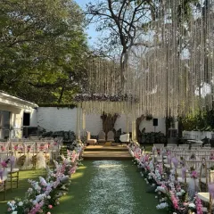 The view as guests arrive. The aisle is lined with abundant pink and white flowers, leading the eye towards the stunning floral mandap nestled under a large tree, creating a natural yet elegant cathedral for the vows.