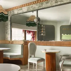 This view shows the interplay of textures and reflections within the cafe. A large, curved mirror expands the space and reflects the custom fringed lighting, while the fluted wood table base and gingham chairs add layers of detail.