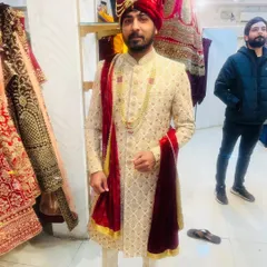 A groom trying on a cream-colored sherwani with a matching red velvet pagri and stole. This is a classic combination for a traditional wedding ceremony.