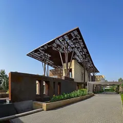 The entrance to the Tree House, where a corten steel wall guides visitors toward the main structure. The driveway approach offers a full view of the dramatic, wing-like roof and its tree-like supports.