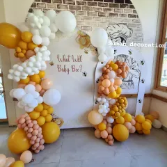 A creative "What Will Baby Bee?" themed gender reveal or baby shower. The setup features a custom backdrop and an organic balloon garland in shades of yellow, orange, and white.