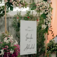 This welcome sign for Sonika and Abhishek's engagement is framed by an arch of exotic white and pink flowers. It’s a simple yet elegant detail that introduces the aesthetic of the event from the moment guests arrive.