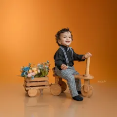 A dapper little boy in plaid pants and a bow tie enjoys a ride on a wooden tricycle. The attached cart full of flowers adds a whimsical touch to this charming studio portrait.