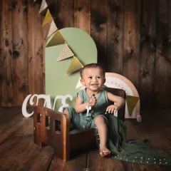 A happy one-year-old boy celebrating his first birthday. This rustic-themed studio setup features a wooden backdrop, a miniature bed, and 'ONE' sign props.
