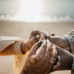 An intimate, close-up photo of the ring exchange during a beach engagement ceremony, with the sparkling ocean in the background.