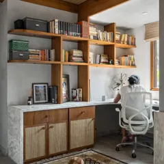 A home office designed for focus and comfort. The custom wooden desk and shelving unit provide ample storage, while the material palette of concrete, wood, and cane creates a calming work environment.