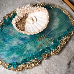 For the couple that loves the sea, I designed this unique ocean-themed ring platter. It features swirling teal and blue resin that mimics ocean waves, a real seashell ring holder adorned with pearls, and a border of sand and crystals. I can personalize it with your initials and special date.
