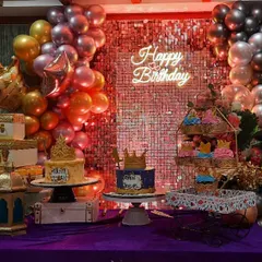 A royal-themed dessert table with a red and silver sequin backdrop. The setup includes multiple cakes with crown toppers, cupcakes, and gold accents for a luxurious feel.