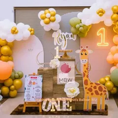 A cheerful and modern first birthday setup with a friendly giraffe character. The design uses a warm color palette of olive green, orange, and gold, complemented by a neon "1" and a personalized milestone board.