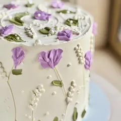 A delicate floral cake featuring hand-piped purple flowers and greenery on a white buttercream base. Tiny edible pearls add a finishing touch of elegance.