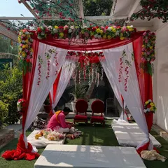 A beautiful mandap for an outdoor wedding, created with red and white drapes and a colorful floral top. The setup includes seating for the pandit and the couple.