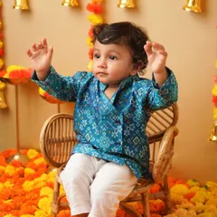 A candid shot of a toddler in a blue kurta, sitting on a chair and raising his hands playfully. This image captures the fun and spirit of a festive photoshoot.