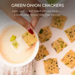 A hand dipping one of my Green Onion Crackers into a creamy dip. Light, savoury, and baked with real herbs, they are a smart and delicious swap for your kids' chip cravings.