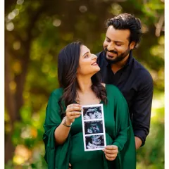 A happy couple shares their excitement in a beautiful park setting. The mother-to-be, dressed in a flowing green gown, proudly displays their baby's sonogram pictures for a lovely pregnancy announcement photo.