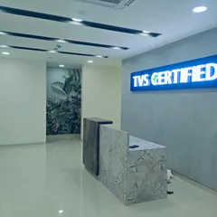The reception desk at the TVS Certified center is crafted from contrasting marble-finish laminates. The backlit "TVS Certified" sign and concrete-texture wall create a modern and trustworthy brand presence.