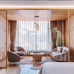 The sitting area in this hotel suite is defined by elegant, art deco-inspired metal screens that separate it from the sleeping area. This creates a multi-functional space that feels both open and private, enhanced by warm wood ceilings and soft lighting.