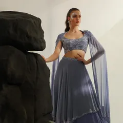 An ethereal look featuring a lavender lehenga with a cape. The styling includes subtle facial glitter, and the pose against the dark rocks creates a beautiful contrast between soft and hard elements.