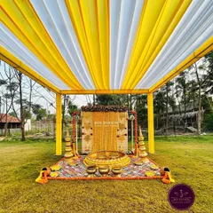 A complete view of an outdoor Haldi setup. The yellow and white draped canopy, the marigold backdrop, and the traditional brass vessels create a picture-perfect scene for the ceremony.