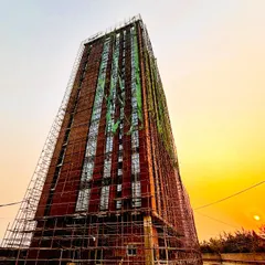 A real-time site photograph of a high-rise building under construction at sunset. The scaffolding creates a temporary exoskeleton around the permanent brick facade, capturing a fleeting moment in the building's creation.