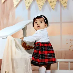 A baby girl in a plaid dress stands by a miniature piano, looking up with a curious and sweet expression.