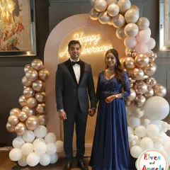 A sophisticated anniversary celebration. The couple is framed by a beautiful balloon arch in rose gold, gold, and white, with a neon 'Happy Anniversary' sign adding a modern touch.