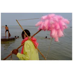 A candy floss seller walks along the banks of the Ganga in Varanasi. The bright pink of the candy contrasts with the earthy tones of the river and the people bathing in the background.