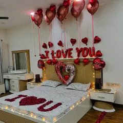 A close-up of the bed decoration, featuring "I LOVE YOU" spelled out with rose petals, heart-shaped foil balloons, and a canopy of red chrome balloons. The warm fairy lights around the headboard add a soft, romantic glow.