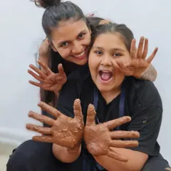Two friends show off their clay-covered hands with excitement. Our workshops are a fantastic group activity, filled with laughter, creativity, and the fun of making something together.