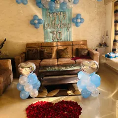 The complete view of the 70th birthday room decoration. It includes blue and silver balloon arrangements, foil 'Happy Birthday' letters, and a beautiful heart made from red rose petals on the floor with initials.