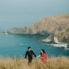 A couple walks hand-in-hand through tall grass on a cliff overlooking the sea. The vibrant colors of their outfits against the natural landscape create a stunning, romantic image.