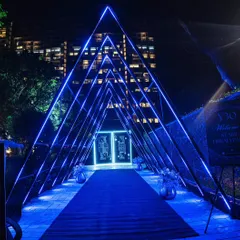 A futuristic and dramatic entrance tunnel made of blue neon triangles, leading to a door with illuminated playing card motifs, setting the stage for a casino-themed Diwali party.