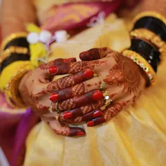 A close-up of a bride's hands, showing her red nail polish, henna design, and traditional bangles.