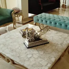 A detailed shot of a living room featuring a custom marble-top coffee table. In the background, you can see a plush green velvet armchair and a tufted turquoise bench, demonstrating our use of rich textures and materials.