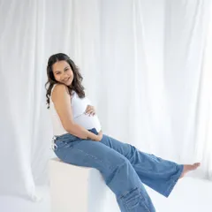 A relaxed and happy portrait. Sitting comfortably in jeans and a tank top, this mom-to-be radiates a calm and confident energy against a simple white backdrop.