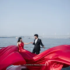 A wide shot of a couple on a yacht in Goa, the massive red trail of her gown spread across the deck, with the sea and bridge in the background.