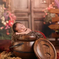 Using unique props can create magical images. Here, a newborn sleeps peacefully atop a rustic wooden pot, surrounded by a soft, floral backdrop that adds a touch of enchantment to the portrait.