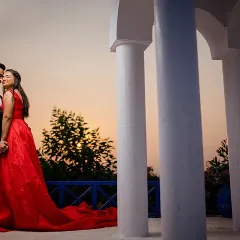 A classic romantic portrait of Yash and Priyanshi at sunset. Her flowing red gown stands out beautifully against the white pillars and the warm colors of the evening sky.