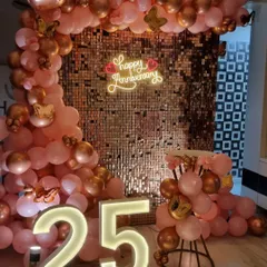A glamorous 25th-anniversary decoration featuring a rose gold sequin wall. The setup is framed by a pink and gold balloon arch and highlighted by large illuminated "25" numbers.
