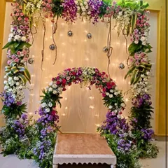 A whimsical and romantic Ganpati setup featuring a double arch of fresh flowers in shades of purple and pink. The backdrop of fairy lights and hanging disco balls adds a touch of playful magic.