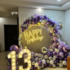 A chic 13th birthday party decoration for a teenager's celebration at home. The decor includes a stunning balloon arch in purple, silver, and white, a "Happy Birthday" neon sign, and marquee numbers.